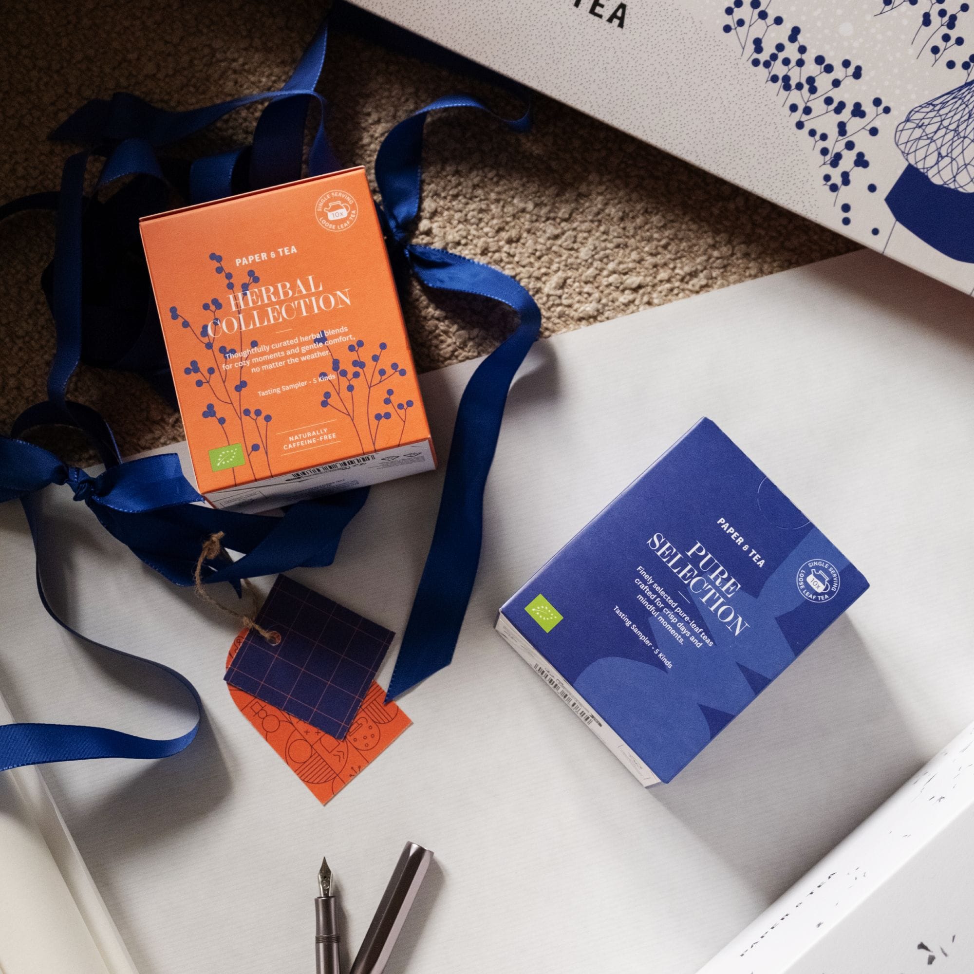 Zwei Paper & Tea Probierboxen – Herbal Collection und Pure Selection – auf weißem Untergrund mit blauem Band und Chrom-Füllfederhalter