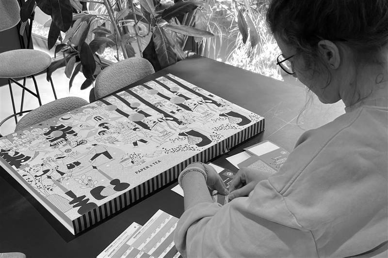 Sylvia rrangiert bunte Verpackungen aus Papier neben einer P&T Tee-Box in einem gemütlichen Raum