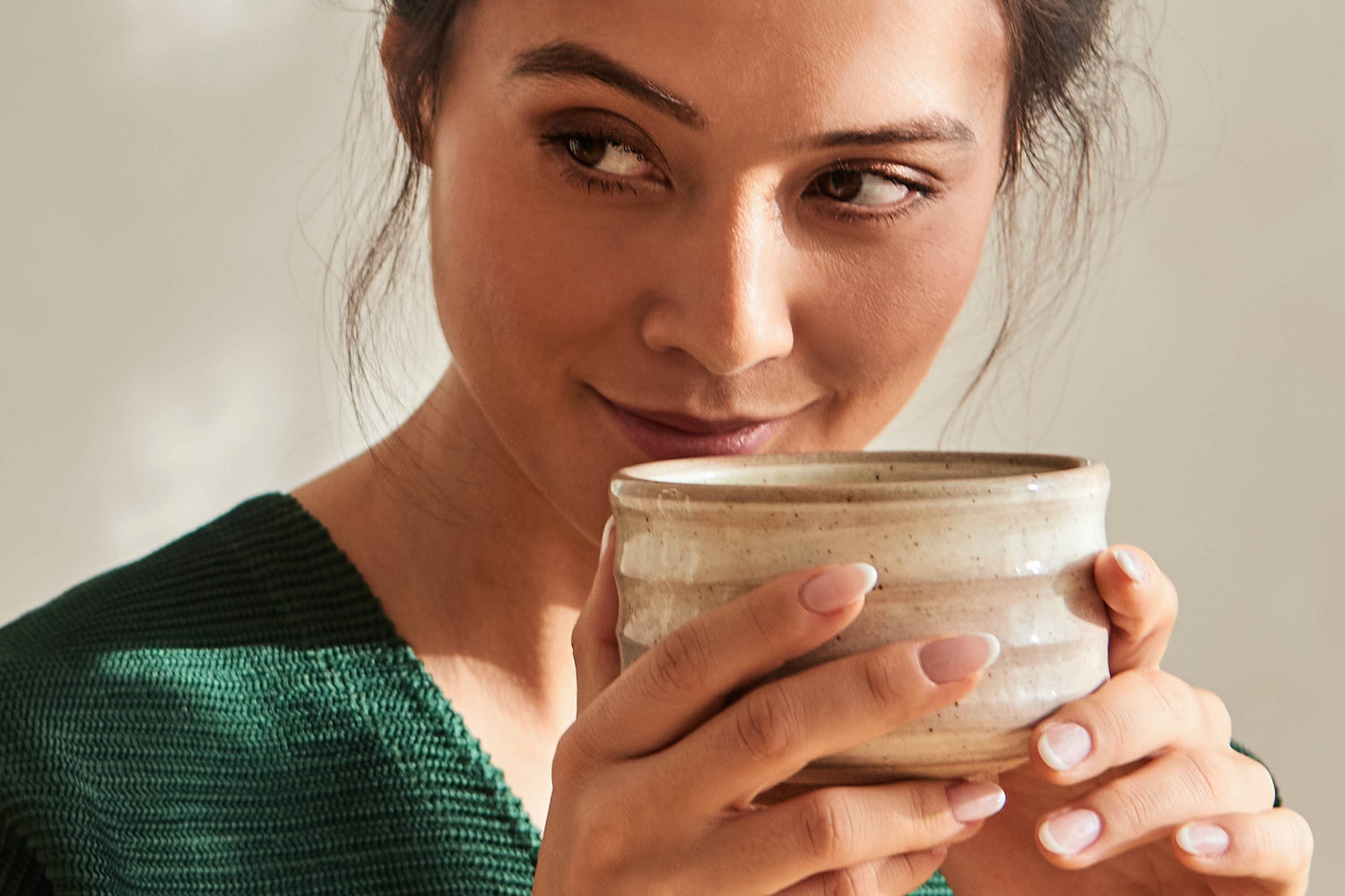 Junge Frau in grünem Strickpullover hält beige Keramiktasse