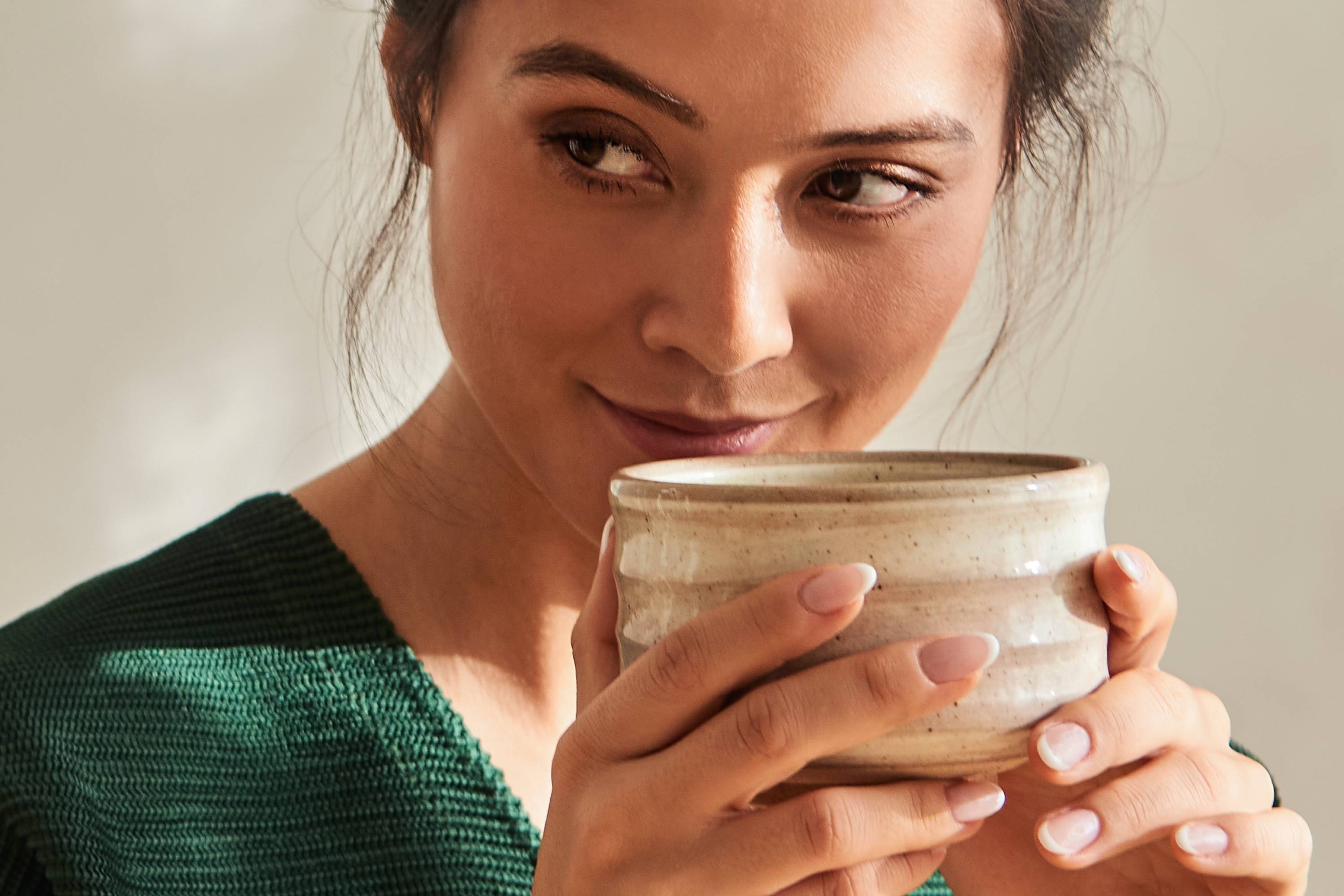 Junge Frau in grünem Strickpullover hält beige Keramiktasse