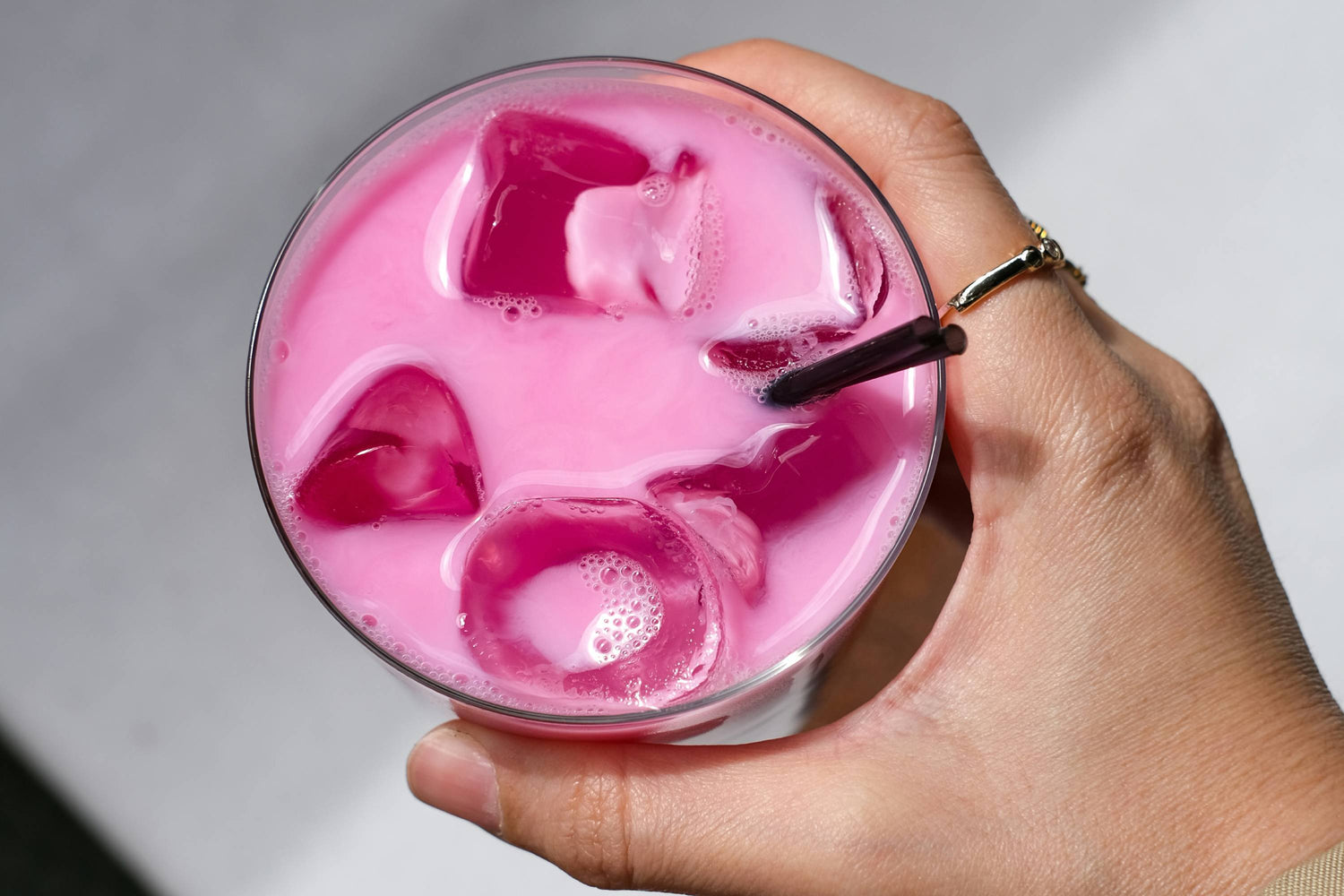 Hand mit Goldring hält Glas mit rosa Getränk und schwarzem Strohhalm
