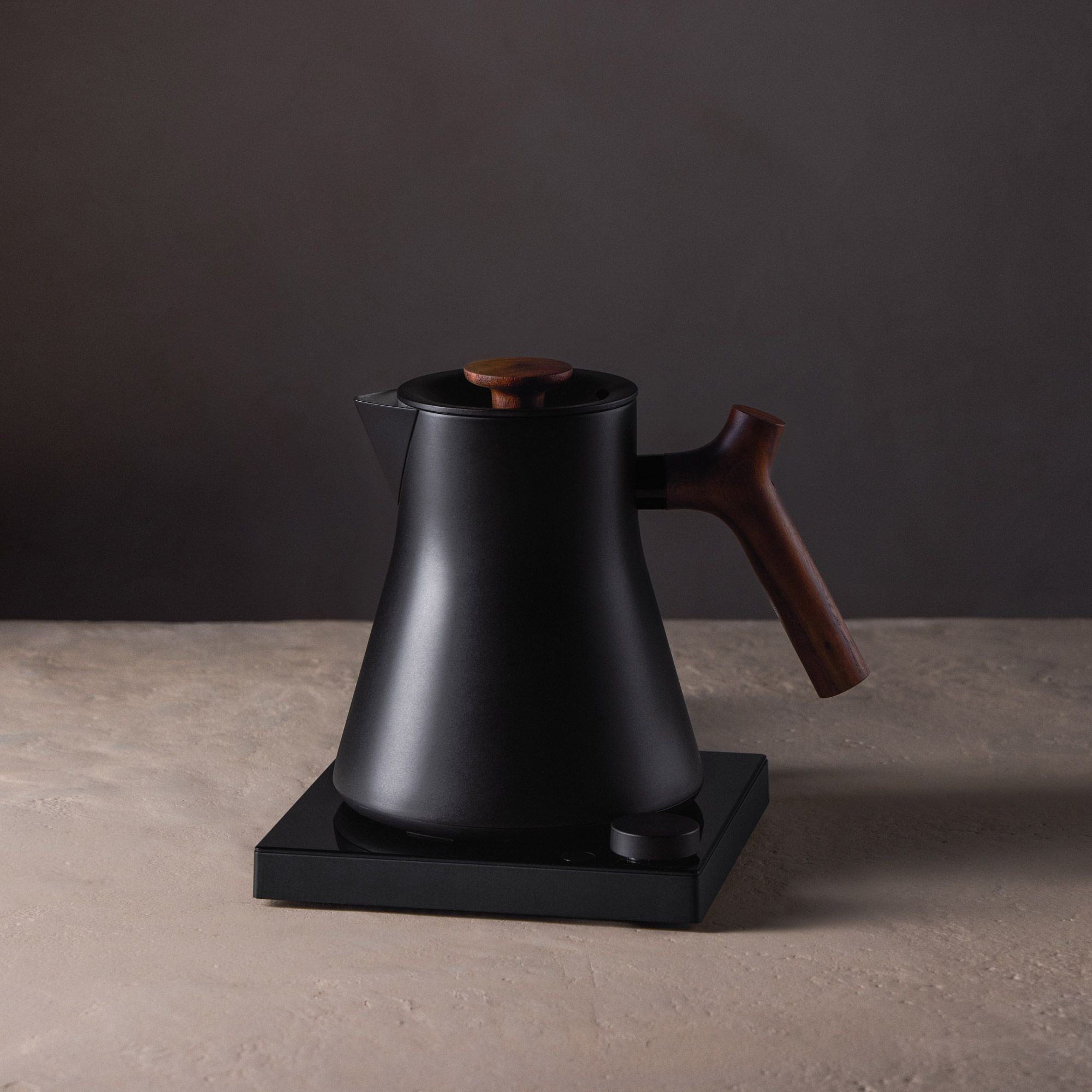 Schwarzer elektrischer Wasserkocher mit Holzgriff auf quadratischem Heizsockel vor dunklem Hintergrund.