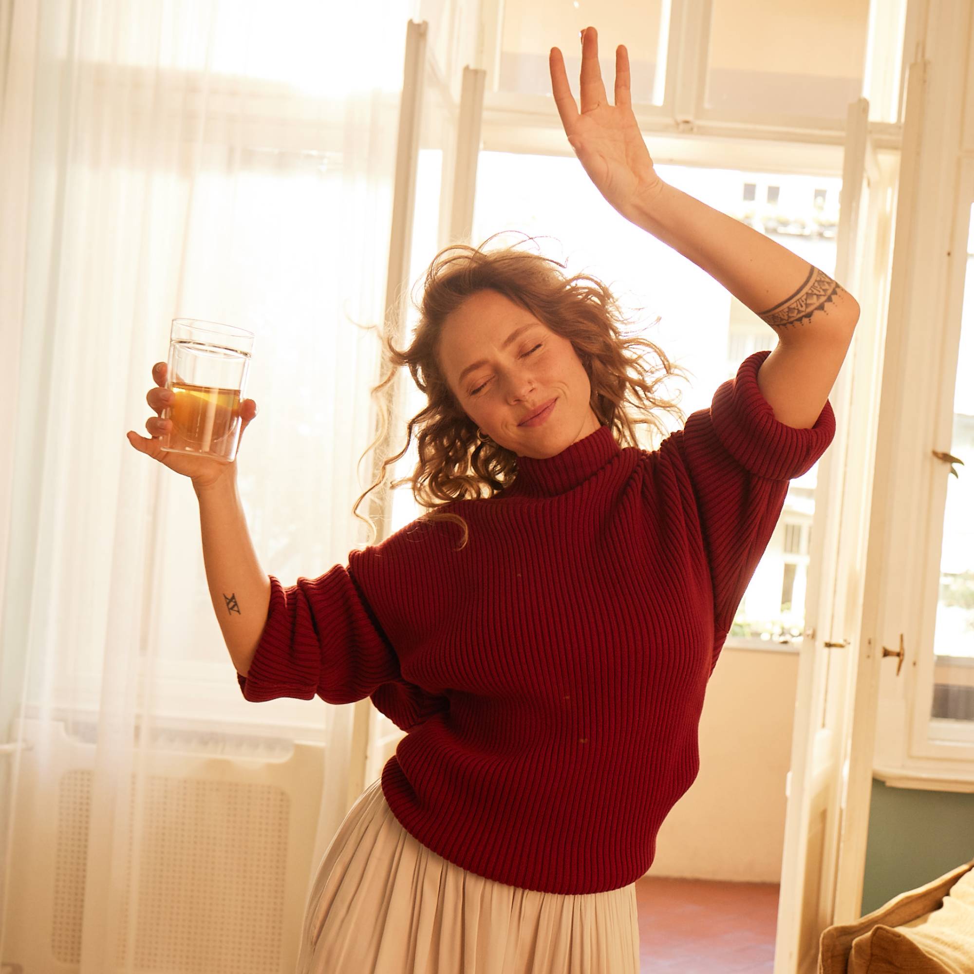Fröhliche Frau in rotem Pullover und beigem Rock tanzt mit einem Glas Tee in sonnigem Raum