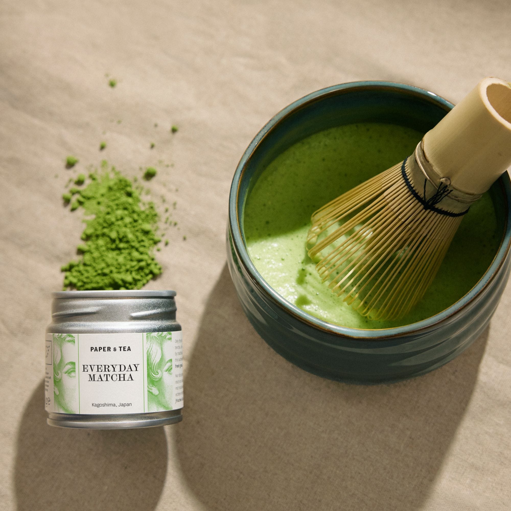 Dose mit grünem Papier und Tee Everyday Matcha aus Kagoshima, neben blauem Keramikbecher mit schaumigem Matcha und Bambusbesen auf beigem Hintergrund.