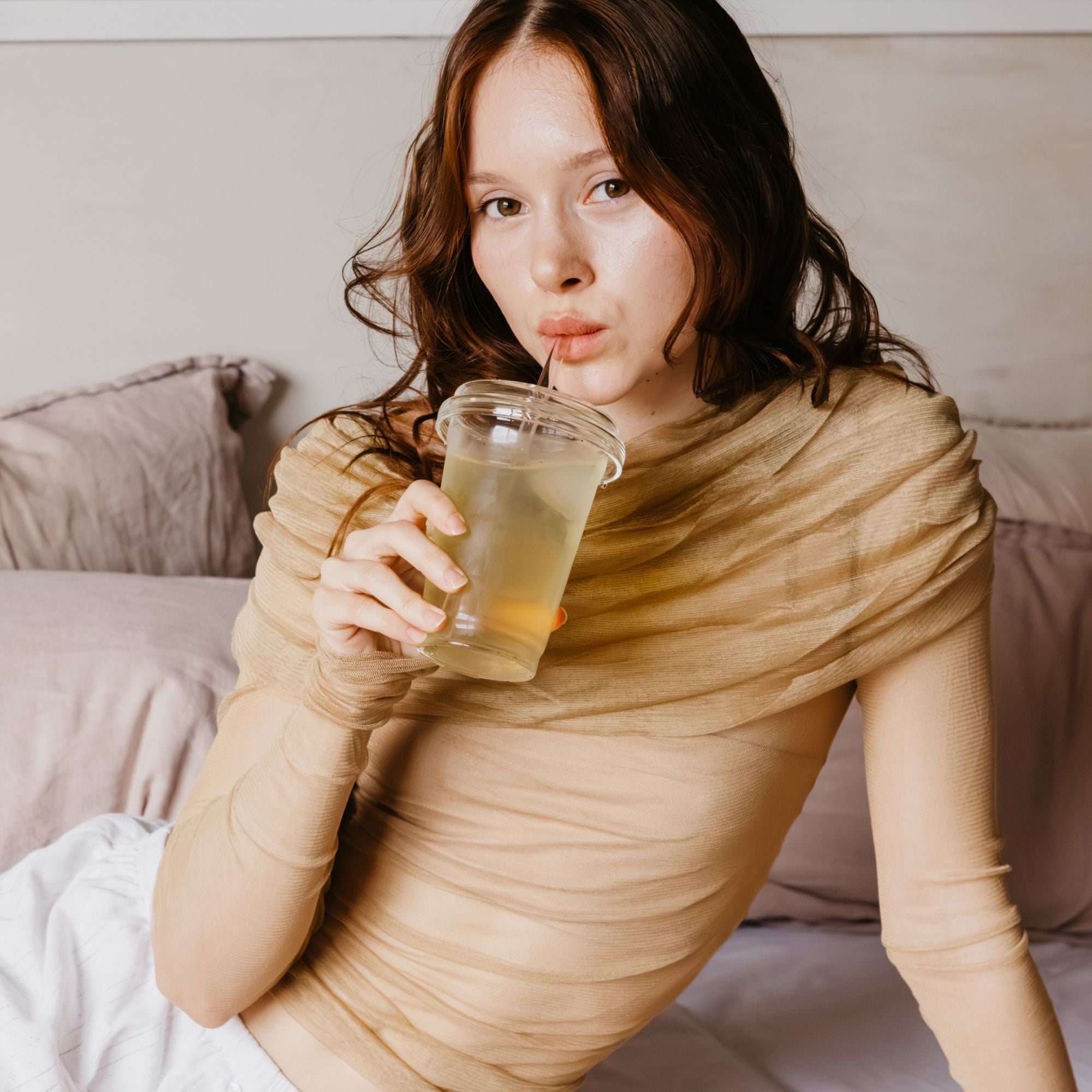 Junge Frau mit braunen Locken trinkt hellgelben Tee aus klarem Becher auf Bett mit rosa und violetten Kissen.