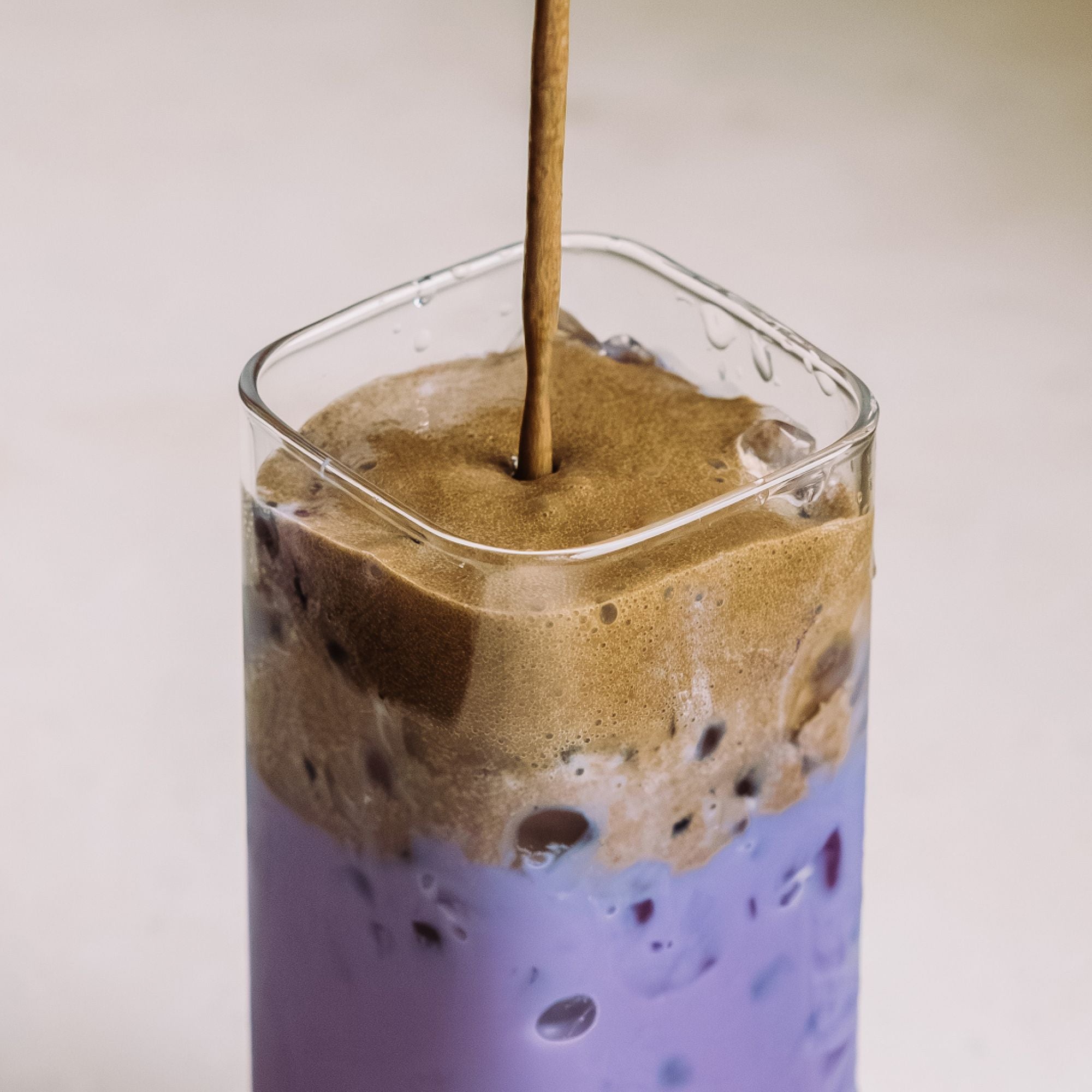 Glas mit violettem Eistee und schaumiger brauner Haube, mit Eiswürfeln, vor hellem Hintergrund