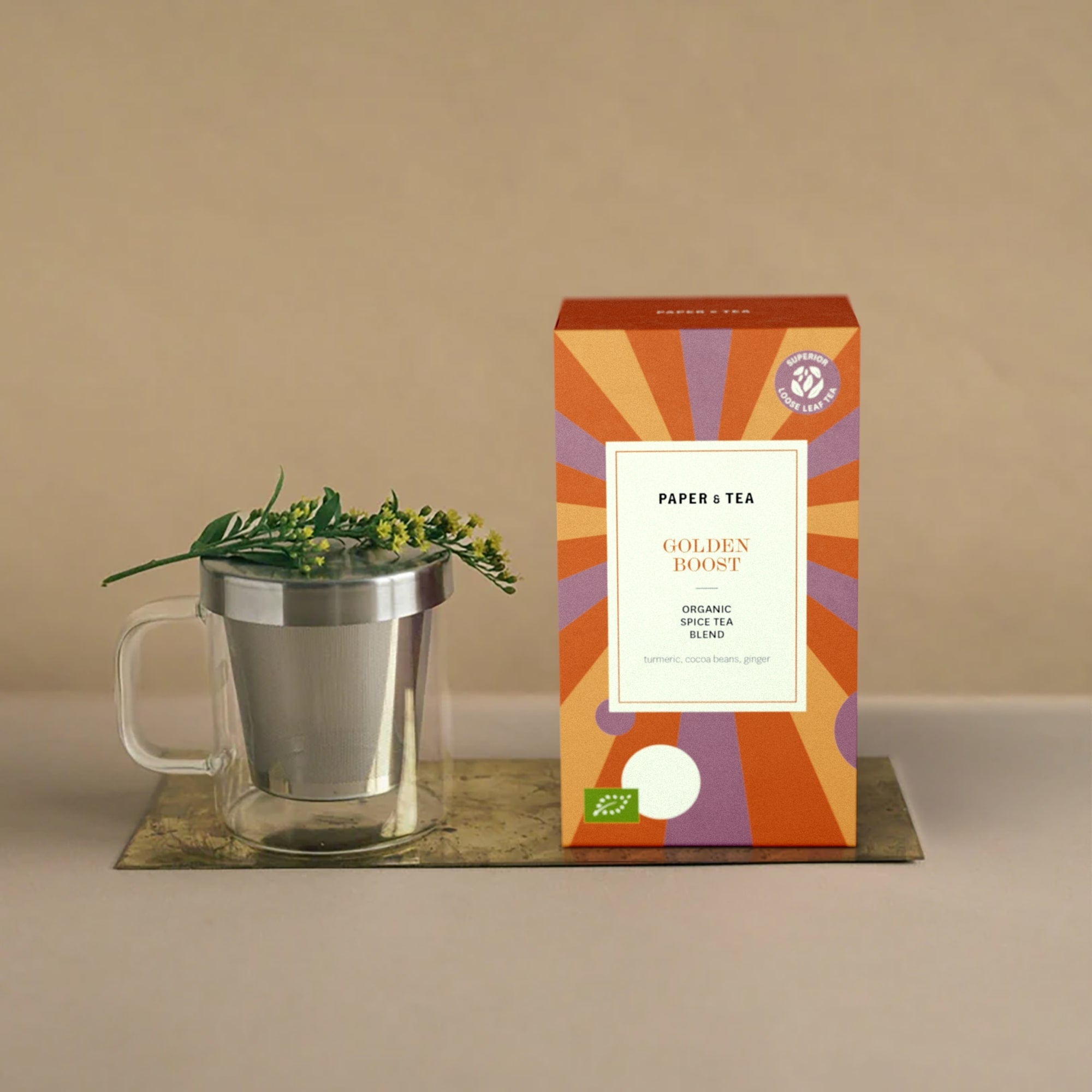 Lebendige GOLDEN BOOST Gewürztee-Verpackung mit Kurkuma, Kakaobohnen, Ingwer und Glasbecher auf Beigeton.