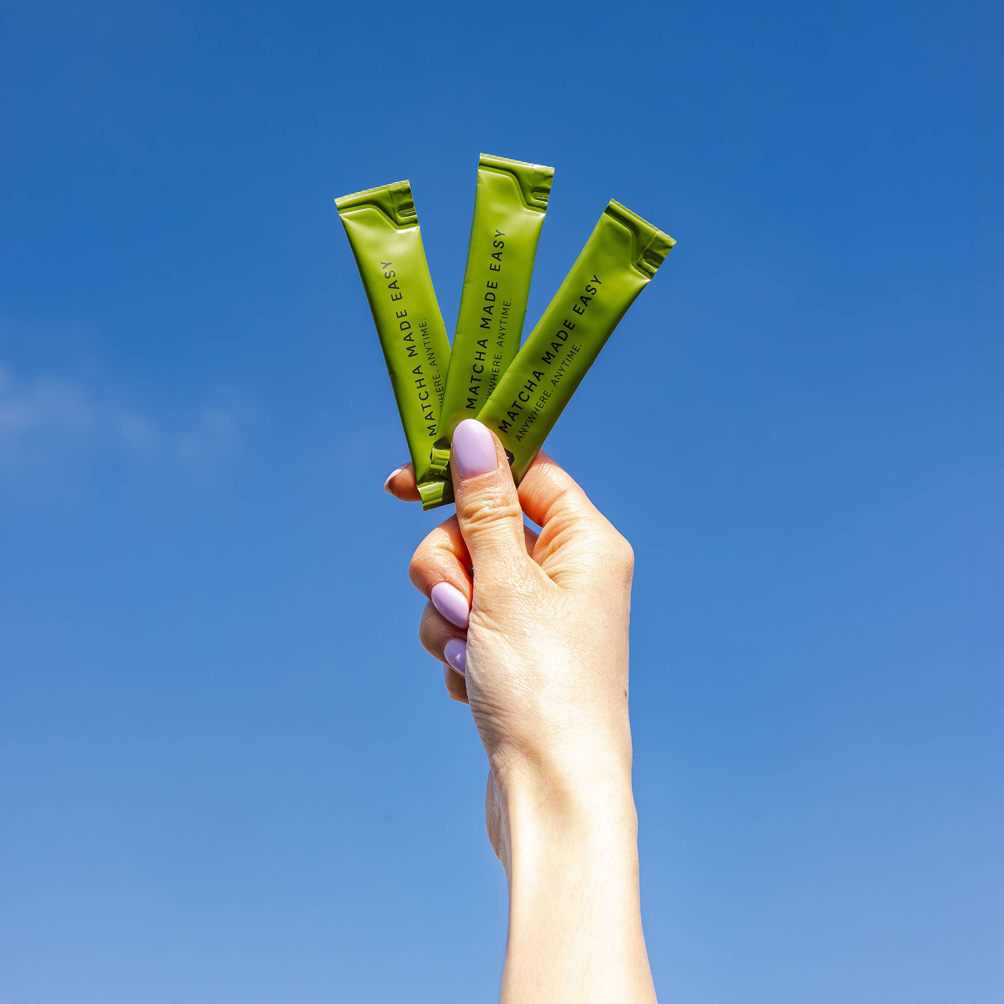 Hand mit lavendellackierten Nägeln hält drei grüne MATCHA MADE EASY-Packungen vor blauem Himmel.