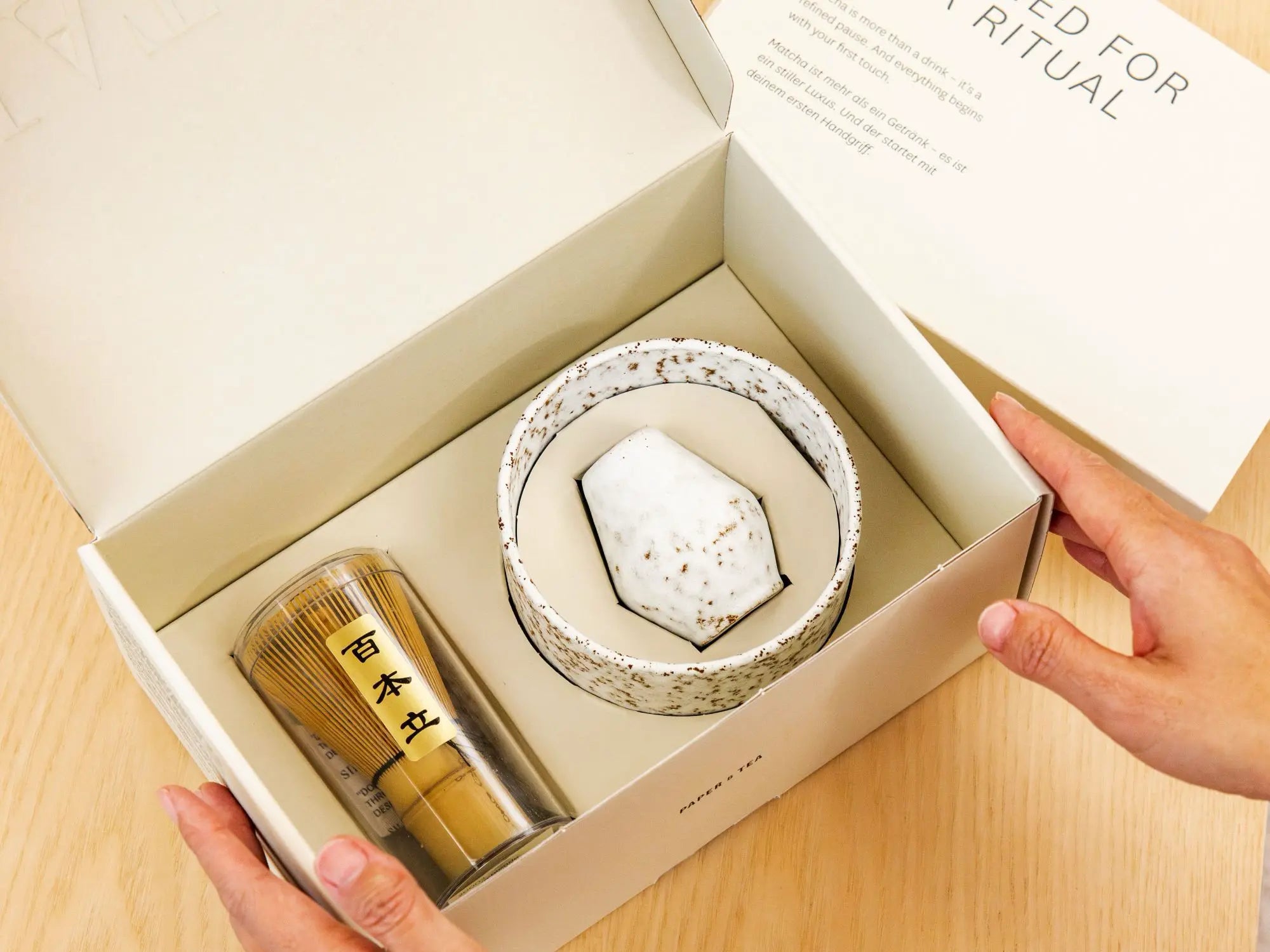 Beige Geschenkbox mit gesprenkelter Keramikschale, Keramikstück und Bambus-Matcha-Besen auf Holztisch.