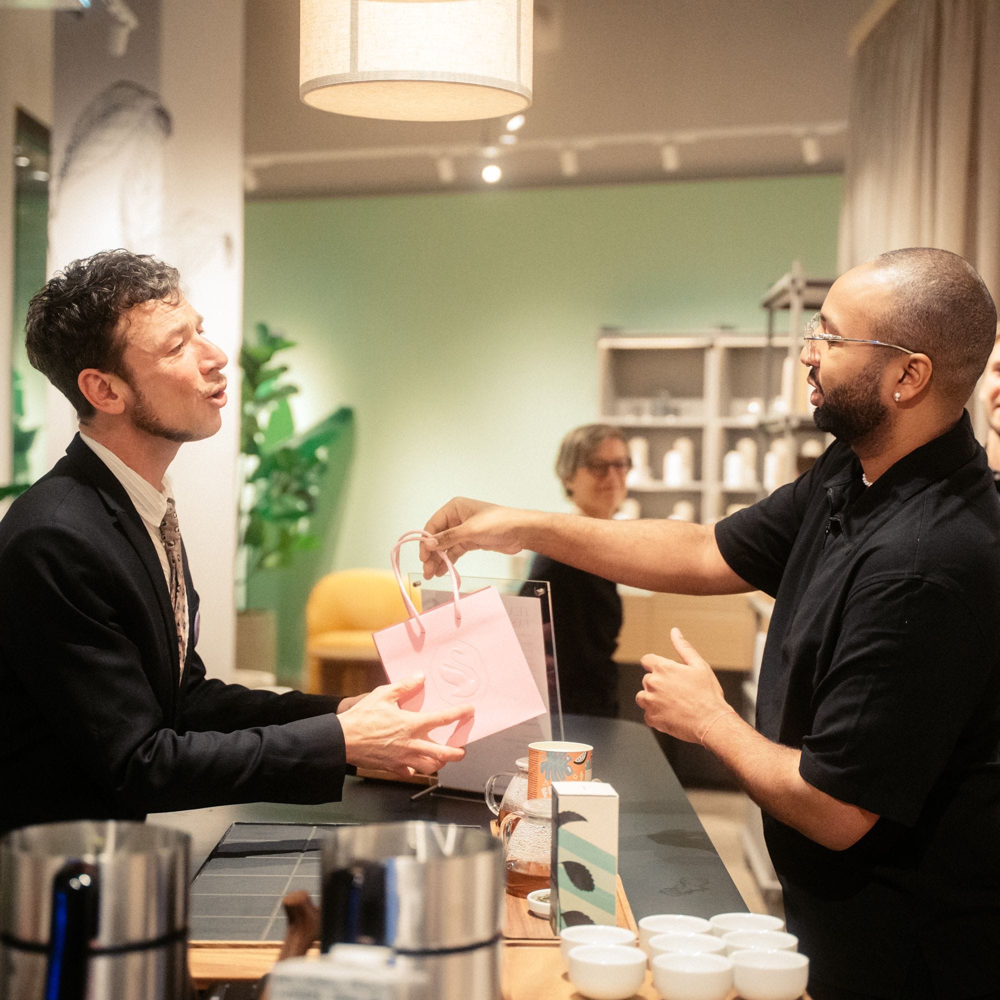 Geschäftsmann erhält pinke Einkaufstasche von Mitarbeiter in modernem, grün gestrichenem Teegeschäft.