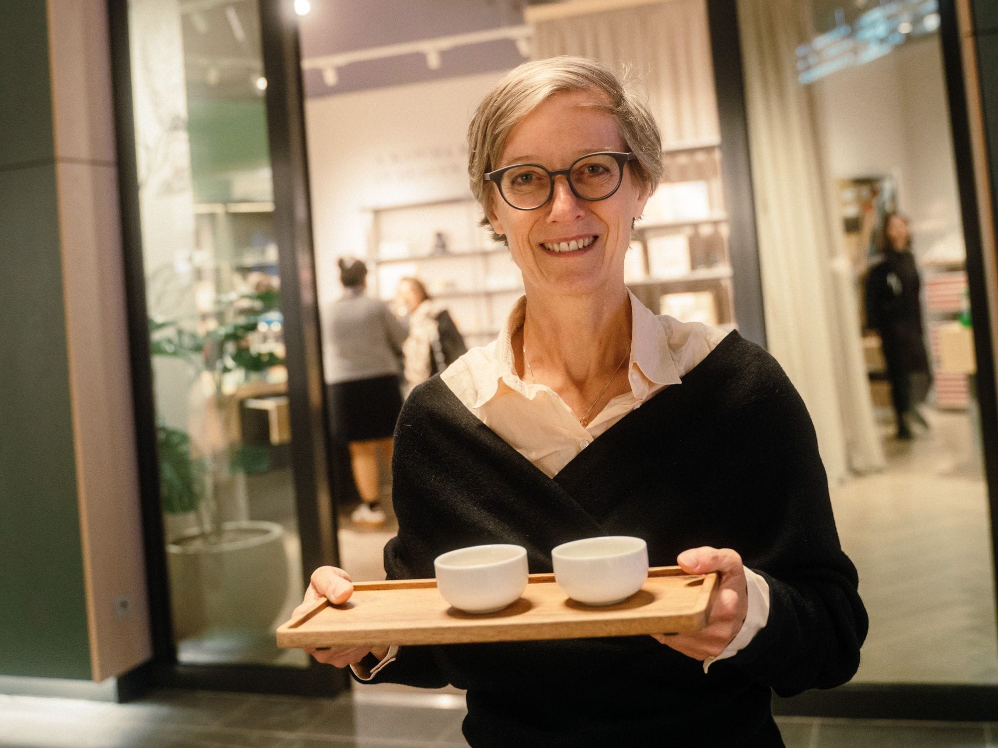 Lächelnde Frau mit kurzem blondem Haar und Brille, hält Holztablett mit zwei weißen Keramiktassen im Café.