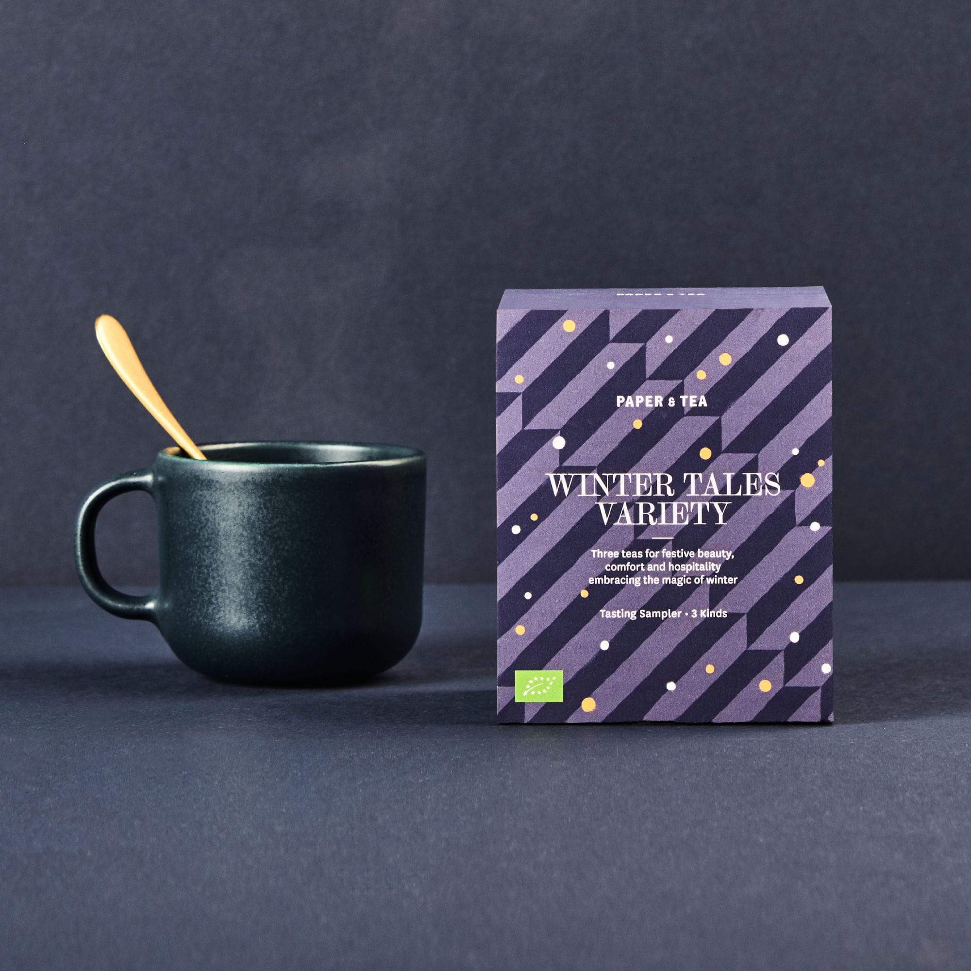 Dunkle Keramiktasse mit goldenem Löffel neben lila Verpackung von PAPER & TEA Winter Tales Teeproben.