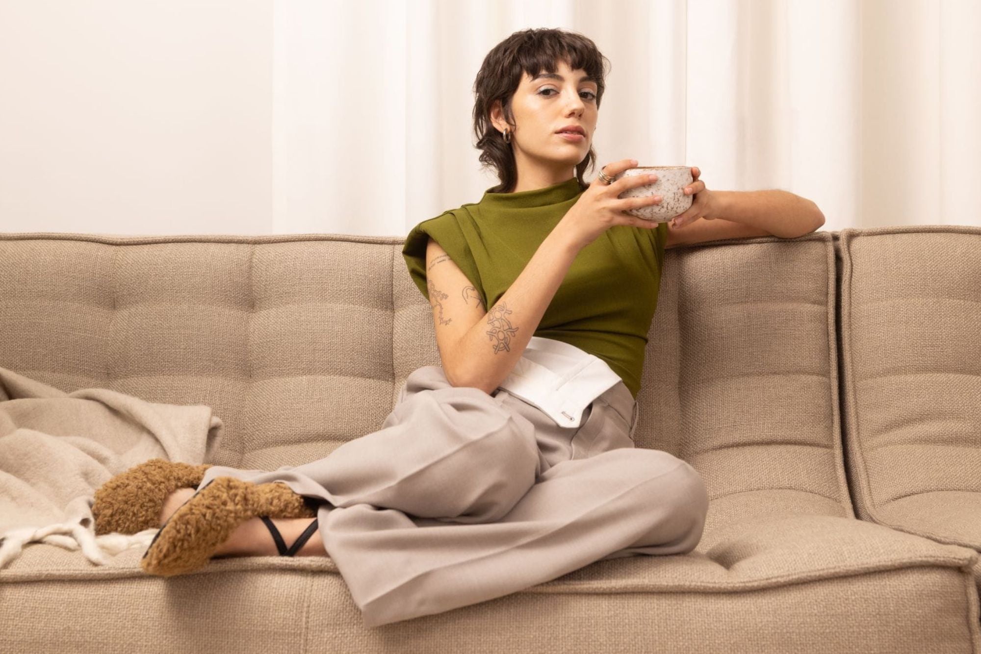 Junge Frau in grünem Top und beiger Hose auf Sofa mit getupfter Keramiktasse nah am Gesicht