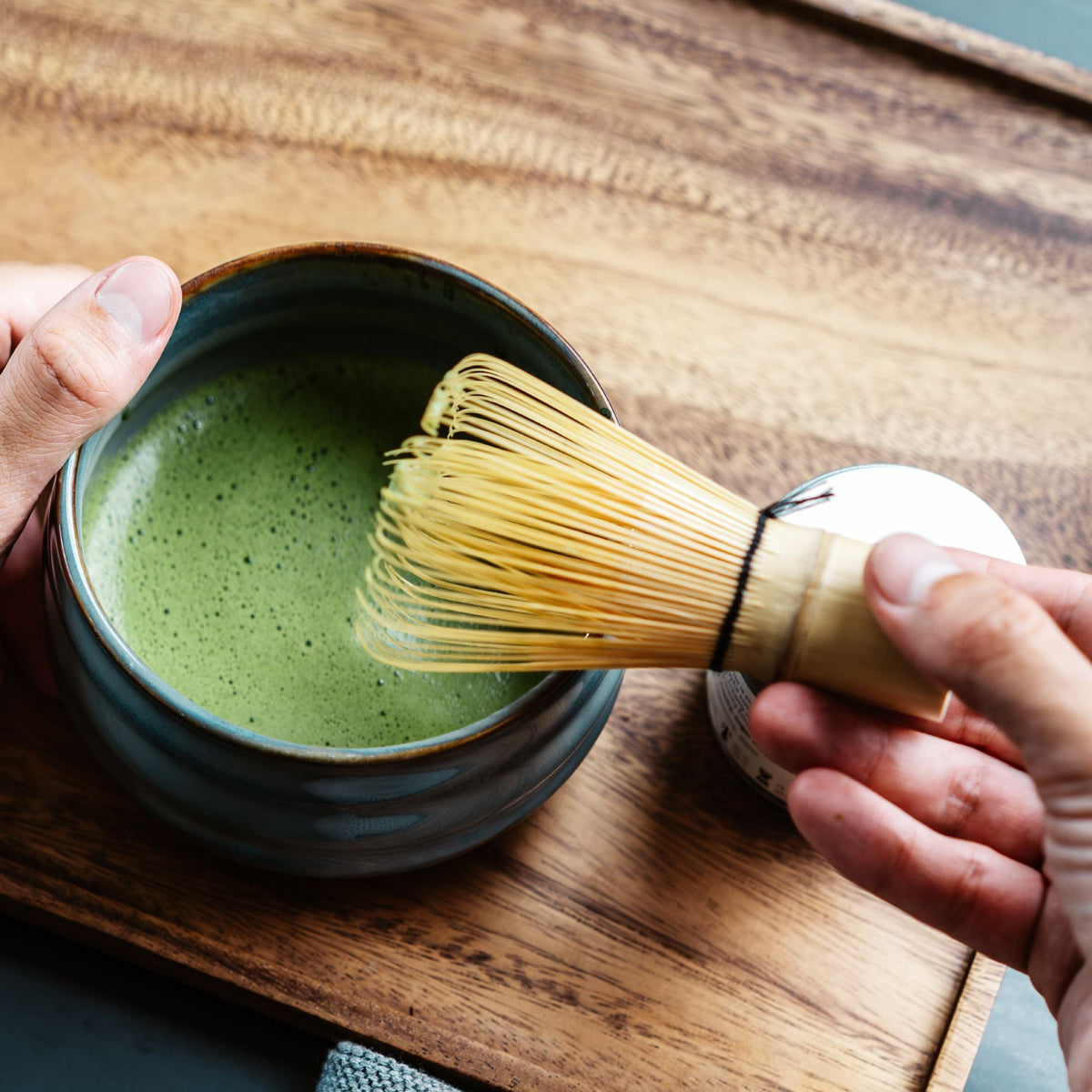 Hände bereiten Matcha-Tee mit Bambusbesen im dunkelgrünen Keramikbecher auf einem Holztablett zu.