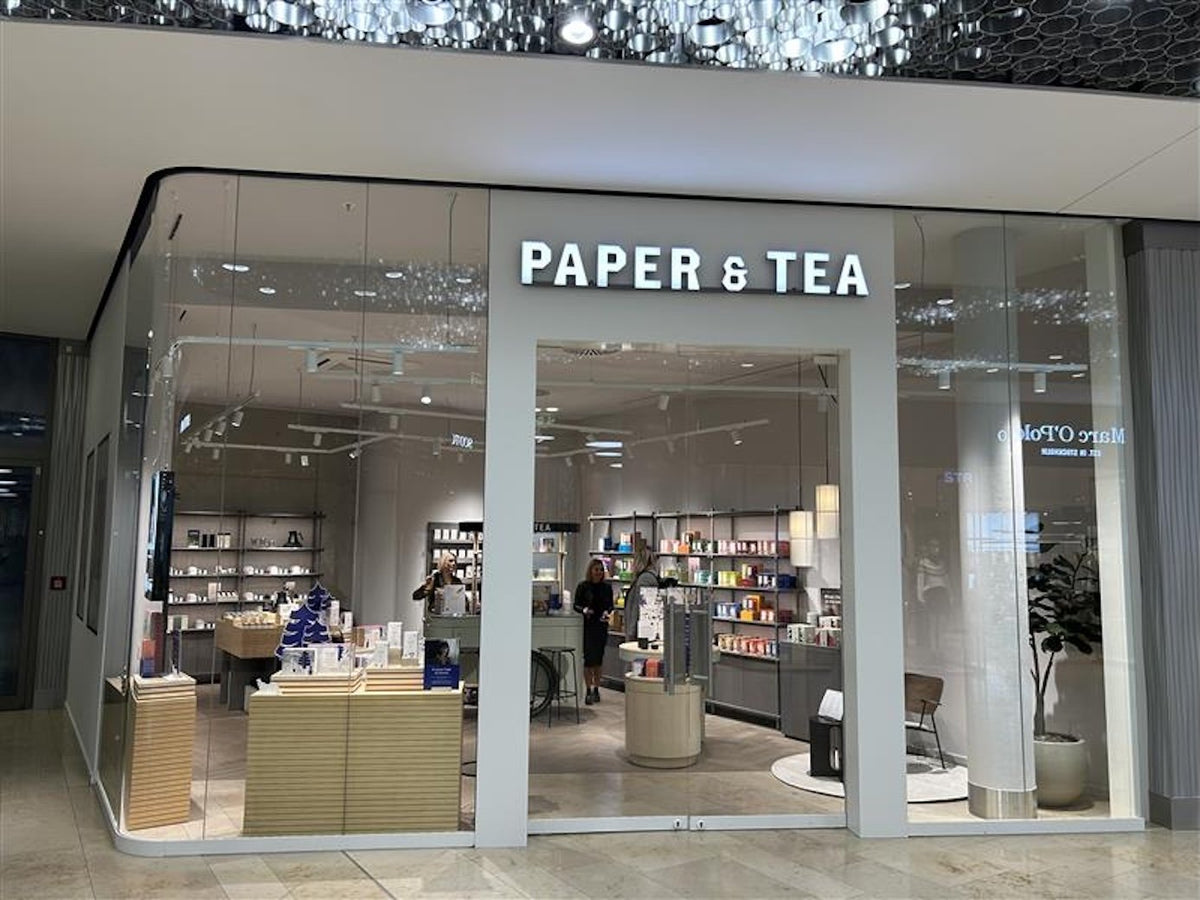 Moderner PAPER & TEA Laden mit Glasfront, beleuchtetem Schild und bunten Teeregale.