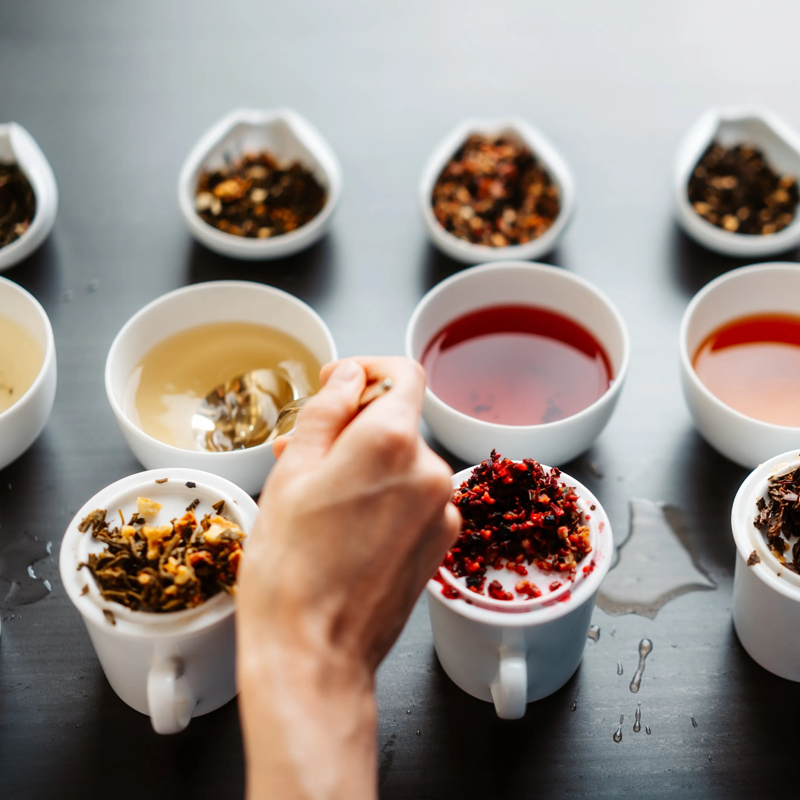 Hand hält Löffel mit roten Teeblättern über weißer Tasse, daneben drei Tassen mit losen Teeblättern.